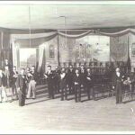 Image: a scattering of men in a large room dressed with lines of chairs and featuring a small stage with a piano.