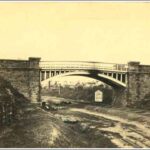 Image: a metal arched bridge with stone abutments passes over a railway track.