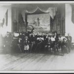 Image: members of a small band, both male and female, pose with their instruments in front of a small stage dressed with a backdrop featuring a castle.