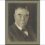 Image: A photographic portrait of a moustachioed middle-aged Caucasian man wearing wire-rimmed spectacles and a mid-20th century suit and tie