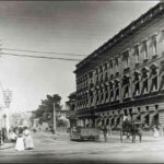 Image: a leisurely scene, with an open horse tram trundling along the road, whilst ladies walk holding sun umbrellas. Two of the ladies are tending to a child in a perambulator. On the right is Eagle Chambers, an imposing three storey building.