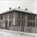 Image: a square two storey stone building with protruding columned portico, shuttered windows and a tiled roof.