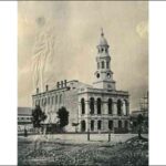 Image: A large stone building with a central tower capped by a dome. A dirt street is visible in the foreground