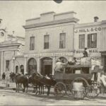 Image: A rectangular single-storey building with central clock tower and cupola is located next to a row of rectangular buildings. A horse-drawn carriage stands in the dirt street in front of these buildings