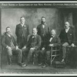 Image: black and white shot of six men, four are sitting and two are standing