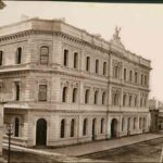 Image: a three storey stone building with corner entrance, arched windows on its ground and third floor and rectangular ones on its second, and a solid parapet with decorative feature in in the centre of one facade.