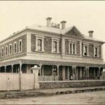 Image: A large, two-storey stone building bounded by a white fence and gate