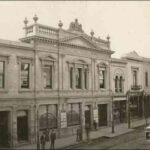 Image: a two storey theatre building, part of a longer terrace, with a decorative parapet featuring stone urns, a lion and a unicorn