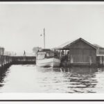 Image: A wooden motor boat with large cabin is moored alongside a jetty with a covered shelter. Several people are transiting from the jetty to the boat