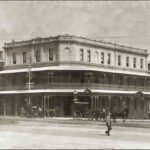 Image: a three storey corner hotel with a protruding balcony on the second floor which also forms a verandah. Horse drawn vehicles pass on the street outside.