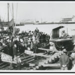 Image: A small motor vessel is launched from its construction slip into a river. A crowd of people in late-Victorian attire look on