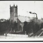 Image: A large stone church building with a tower topped by four spires stands at the corner of two dirt roads. A man stands in front of the churchyard fence