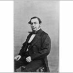 Image: A man wearing mid-Victorian era attire poses for a photograph. He is seated and is wearing a suit and jacket with tails, and has a fob watch on a chain attached to his waistcoat.