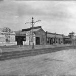 Image: a series of small single storey industrial buildings lining a wide dirt road. Many of the buildings feature painting signs advertising the products sold inside.