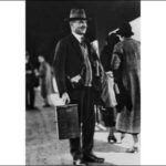 Image: A man in a hat and suit stands on a platform at a railway station. He is carrying a wooden box and several leather equipment satchels