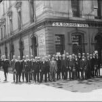 Image: A group of men wearing 1920s era suits and holding their hats pose for a photograph on a street corner outside a building with a sign reading: "S.A. Soldiers' Fund". To one side are two more men who only have one leg.