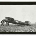 Image: Three men stand next to a First World War-era monoplane parked on a large, flat expanse of ground