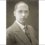 Image: A photographic head-and-shoulders portrait of a young, clean-shaven Caucasian man wearing an early-20th century suit and wire-rimmed spectacles