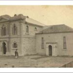 Image: A man in a top-hat and Victorian clothing stands in front of a large, two-storey rectangular stone building