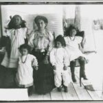 Image: A woman in Edwardian dress sits on a verandah with her four children. A corrugated water tank is visible in the background