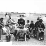 Image: A group of Afghan children and an Aboriginal woman sit around a small table, atop which sits a gramophone. A Caucasian woman in Victorian dress operates the gramophone