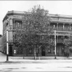 Image: a large tree obscures the view of a three storey stone building on the corner of a wide street and small alley. The building has balconies on its second and third floor and a corner door at street level.