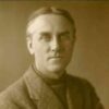 Image: A photographic head-and-shoulders portrait of a clean-shaven man wearing a turtleneck sweater and tweed jacket