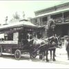 Image: a man in early 20th century clothing stands by a horse drawn tram which is parked in front of a two storey house.