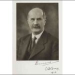 Image: A photographic portrait of a balding, moustachioed middle-aged man wearing a suit and tie