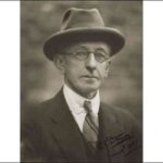 Image: A photographic head-and-shoulders portrait of a clean-shaven middle-aged man. The poet looks directly at the camera through round, wire-rimmed spectacles. He is wearing a collar, tie and jacket and a fedora hat