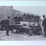 Image: Two men in suits and hats examine a section of aircraft wreckage. A stone house is visible in the immediate background