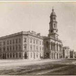 Image: A large stone building with a central tower capped by a dome. A dirt street is visible in the foreground