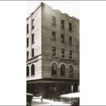 Image: a five storey tower building with shops on its ground floor. Each side has three windows to each floor. On the second storey they are arched while on floors three to five they are rectangular.