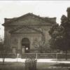 Image: A man wearing a white coat and a peaked cap stands in front of a sandstone building with arches, columns and decorative gables.