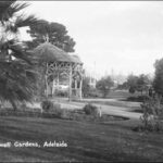 Image: Black and white photograph of a rustic wooden rotunda situated in public gardens. Indistinct figures wander along the garden paths in the middle-ground while buildings can be seen in the distance.