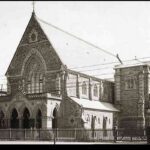 Image: A large bluestone church is surrounded on three sides by a picket fence and borders a road. Electric trolley lines are visible in front of the church