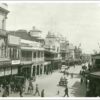 Image: A view of a city street with many people in 1920s attire walking, riding horses or driving motor vehicles. The street is lined with 2 and 3 storey commercial buildings, most with verandahs and/or balconies and a number featuring towers with domes.