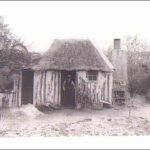 Image: An elderly man stands in the front doorway of a simple, single-storey timber hut. The hut is surrounded by a few scrubby trees