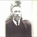 Image: A photographic head-and-shoulders portrait of a middle-aged man with a goatee and full head of hair. He is wearing an Edwardian-era suit and tie