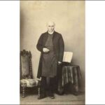 Image: Sepia photograph of bald man standing in front of a chair and table