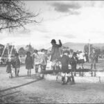 Image: A group of children stand near the edge of a pond in a playground. One child balances on the edge of the pond. A flagpole and playground equipment are visible in the background