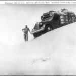 Image: Man stands leaning on heavily loaded truck perched on top of a sandhill