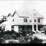 Image: A large, two-storey mansion stands at the top of a hill surrounded by trees and vegetation. A small group of men and women stand in front of the house