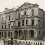 Image: A three storey brick building with a stone façade. The building is of two distinct halves. The right half features an arched porch , a second storey bay window, balconies and a decorative pediment. The left is plainer and features a simple parapet.