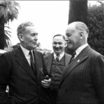Image: Three men in suits and ties speak with one another on a property's well-manicured grounds. Each man is smiling, suggested he is amused by the conversation