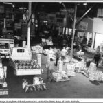 Image: men and women unload boxes of fruit and vegetables from 1980s era trucks in a covered market with shops lining one wall