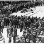 Image: A large group of men wearing suits and hats congregate near a train line