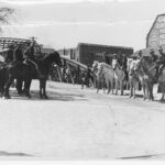 Image: Several men on horseback form two ranks in front of an iron bridge