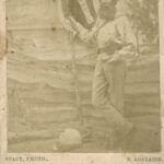 Image: Photograph of bearded man standing in front of a painted background of a beach. He stands in three quarter profile, legs crossed, and holds a flag in his right hand. His hat sits on the ground in front of him.