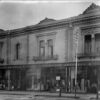 Image: a black and white photograph of a two storey terrace building with large plate glass shop windows on the first floor displaying a range of clothing and fabrics